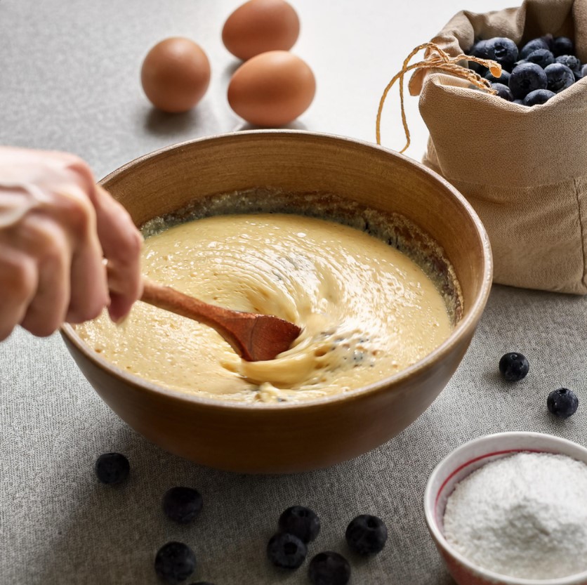 Hand mixing blueberry muffin batter with a wooden spoon in a bowl, blueberries visible.